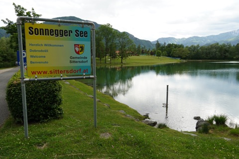 Ufer des Sonnegger Sees mit Begrüßungstafel, Liegewiese und Karawanken im Hintergrund