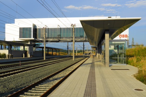Bahnsteig des neuen Bahnhofs Kühnsdorf-Klopeiner See mit Gleisen, Oberleitung, geschwungenem Bahnsteigdach, gläsernem Wartehäuschen und dem weißen Stationsgebäude mit Fußgängersteg über den Gleisen in der Abendsonne