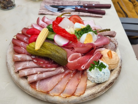 Runde Holzplatte mit Kärntner Jause: Schinken, Speck, Salami, Gurkerl, Paprika, Radieschen, Maiskolben, Ei und zwei Aufstriche, dahinter Besteck auf einer Serviette