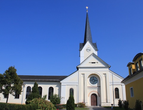 Helle Pfarrkirche von St. Kanzian mit spitzem Turm und Uhr vor blauem Himmel, rechts ein gelbes Haus