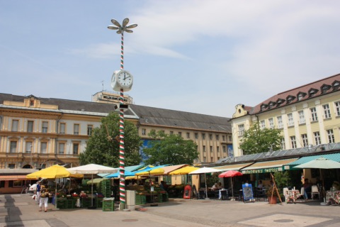 Marktstände mit bunten Sonnenschirmen und die Marktuhr auf dem Benediktinerplatz in Klagenfurt