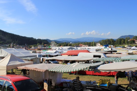 Marktwiese des Bleiburger Wiesenmarkts mit vielen Verkaufsständen und Zelten, im Hintergrund Riesenrad und Fahrgeschäfte, dahinter bewaldete Hügel und Berge unter blauem Himmel, vorne links ein Autodach