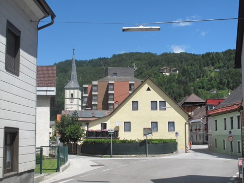 Straße in Bad Eisenkappel mit Wohnhäusern und dem spitzen Kirchturm, dahinter ein bewaldeter Berghang unter blauem Himmel