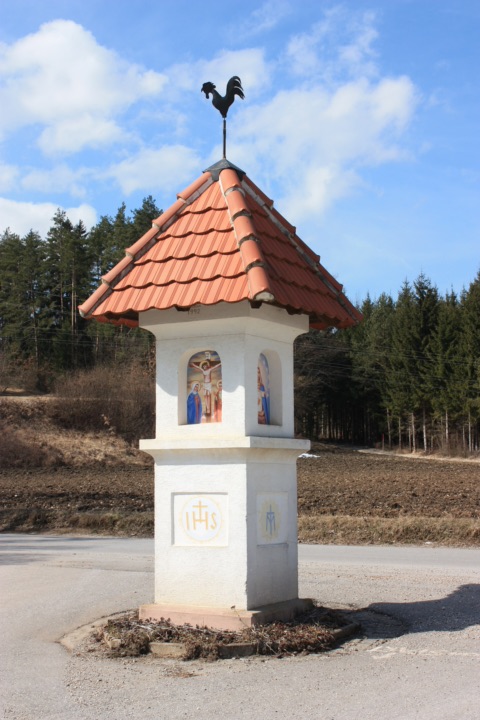 Weißer Bildstock mit rotem Ziegeldach, Wetterhahn und Heiligenbildern an einer Straße in Hof bei Eberndorf, dahinter Acker und Wald