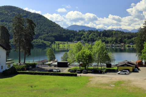 Blick von der Norduferstraße über einen Parkplatz und Bäume auf den Klopeiner See, dahinter ein bewaldeter Hügel und Berge unter Wolken