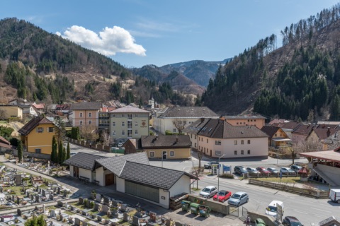 Blick über den Ortskern von Bad Eisenkappel mit Häusern, Friedhof und Kirchturm im Tal, umgeben von bewaldeten Bergen