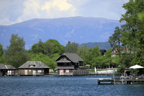Blick vom Südufer über den Klopeiner See auf hölzerne Bootshäuser und einen Badesteg am Nordufer, dahinter die Koralpe