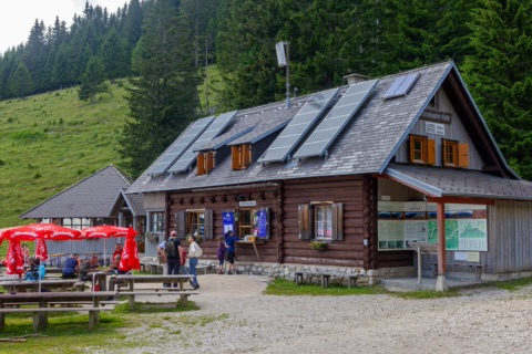 Die Eisenkappler Hütte, ein Blockhaus mit Solarpaneelen auf dem Dach, davor Tische, Bänke und rote Sonnenschirme mit Wanderern, dahinter Wald und Almwiese