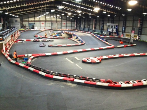 Kurvige Indoor-Kartbahn in einer großen Halle mit rot-weißen Reifenbarrieren, einigen Karts auf der Strecke und einem Streckenposten in Warnweste