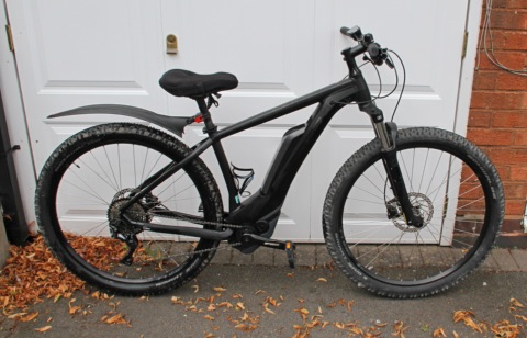 Schwarzes E-Mountainbike mit Akku im Rahmen, abgestellt vor einer weißen Tür, davor Herbstlaub