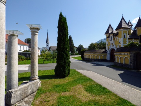 Dorfstraße in Globasnitz mit römischen Säulen, Kirchturm und einem gelben Torbau im Sommer