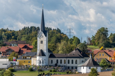Weiße Pfarrkirche von St. Kanzian mit spitzem Kirchturm und Friedhof, umgeben von Häusern und herbstlichen Bäumen