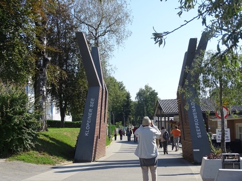 Eingang zur Seepromenade am Klopeiner See mit zwei geschwungenen Stelen mit den Aufschriften „Klopeiner See“ und „Seepromenade“, dahinter Spaziergänger auf dem Weg