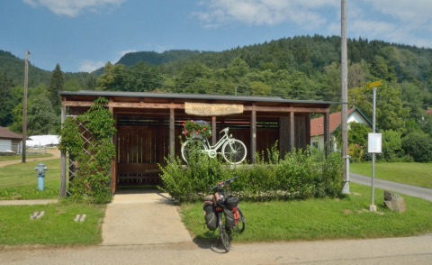 Hölzerner Rad-Rastplatz „Radrast St. Nikolai“ mit Blumenschmuck und einem weißen Deko-Fahrrad, davor ein Reiserad mit Packtaschen, dahinter bewaldete Hügel