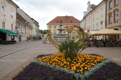 Hauptplatz von St. Veit an der Glan mit Blumenbeet und herzförmiger Skulptur, dahinter ein Brunnen, Bürgerhäuser und Straßencafés