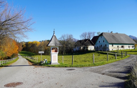 Ortseinfahrt von Lanzendorf mit Bildstock und Ortsschild, dahinter eine eingezäunte Wiese und ein altes Bauernhaus, Herbstlaub unter blauem Himmel