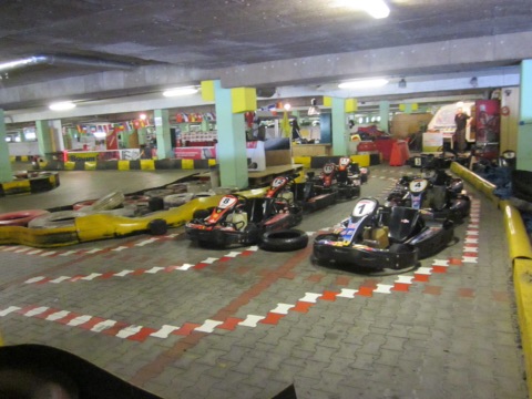 Indoor-Kartbahn in einer Halle: nummerierte Karts stehen hintereinander auf der Strecke mit rot-weißen Fahrbahnmarkierungen, Reifenstapel und gelbe Banden als Begrenzung