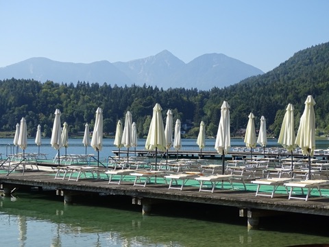 Holzsteg eines Strandbads am Klopeiner See mit Liegen und geschlossenen Sonnenschirmen, dahinter der See, bewaldetes Ufer und der Hochobir