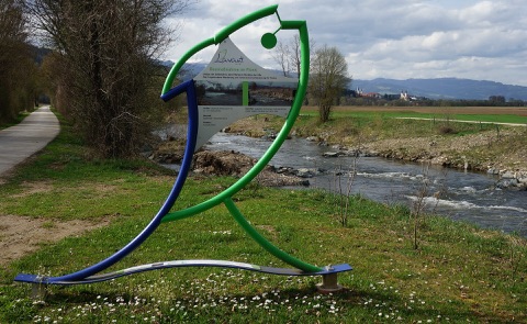 Asphaltierter Weg am Ufer der Lavant bei St. Andrä, davor eine Infoskulptur in Fischform, im Hintergrund Kirchtürme und ein Bergrücken