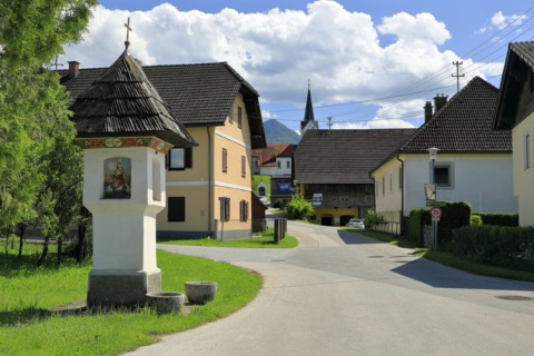 Kirchstraße in Gösselsdorf mit bemaltem Bildstock, Bauernhäusern und Kirchturm vor einem Berg und Sommerwolken
