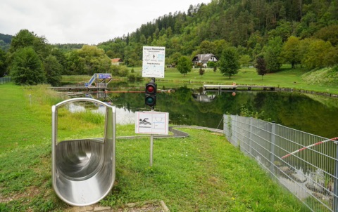 Naturbadesee Lavamünd mit Wasserrutsche, Steg und Liegewiese vor bewaldetem Hang
