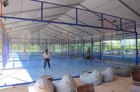 Verglaste Padel-Plätze mit blauem Kunstrasen in einer Zelthalle, auf denen einige Personen spielen, vorne Sitzsäcke