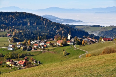 Herbstlicher Blick auf das Dorf Diex mit der Wehrkirche auf dem Sonnenplateau, ringsum Wiesen und Wald, im Hintergrund Nebel im Tal und Bergketten