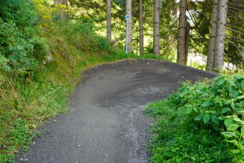 Steilkurve aus Schotter auf einem Mountainbike-Trail im Fichtenwald, daneben ein kleines blaues Streckenschild