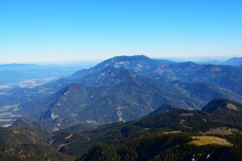 Blick aus der Höhe auf das bewaldete Bergland der Karawanken mit dem breiten Gipfelkamm der Petzen unter klarem blauem Himmel