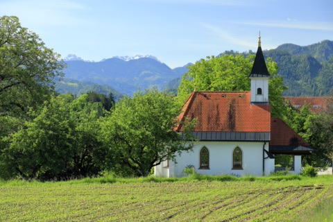 Weiße Kapelle mit rotem Ziegeldach und Türmchen am Rand von Rückersdorf, davor ein Feld, dahinter bewaldete Hügel und schneebedeckte Gipfel der Steiner Alpen