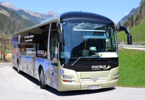 Überlandbus mit Postbus-Schriftzug, geparkt vor einem Alpental mit Bergen