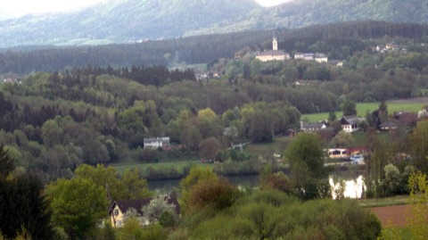 Blick über bewaldete Hügel und die Drau auf Tainach mit Kirche und großem Propsteigebäude auf einer Anhöhe