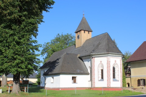 Weiße Pfarrkirche Kühnsdorf mit hölzernem Glockenturm und rot-weiß verzierten Fenstern, daneben eine große Linde, blauer Himmel