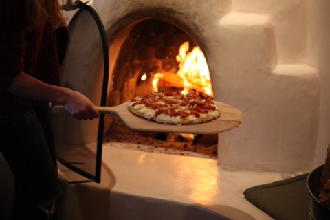 Eine Pizza mit Tomaten und Mozzarella wird auf einem Holzschieber vor einem lodernden Holzofen gehalten