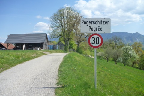 Zweisprachige Ortstafel Pogerschitzen – Pogrče mit 30er-Schild an einer schmalen Dorfstraße, daneben ein Holzstadel, blühende Obstbäume und Berge im Hintergrund
