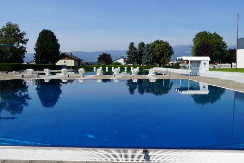 Großes blaues Schwimmbecken im Stadionbad Wolfsberg mit Startblöcken, dahinter Bäume und die Hügel des Lavanttals