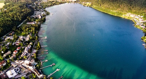 Luftaufnahme des Klopeiner Sees mit türkisgrünem, klarem Wasser am Ufer, vielen Badestegen und Uferbebauung, dahinter Wald