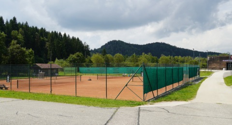 Eingezäunte Tennis-Sandplätze vor bewaldeten Hügeln unter bewölktem Himmel