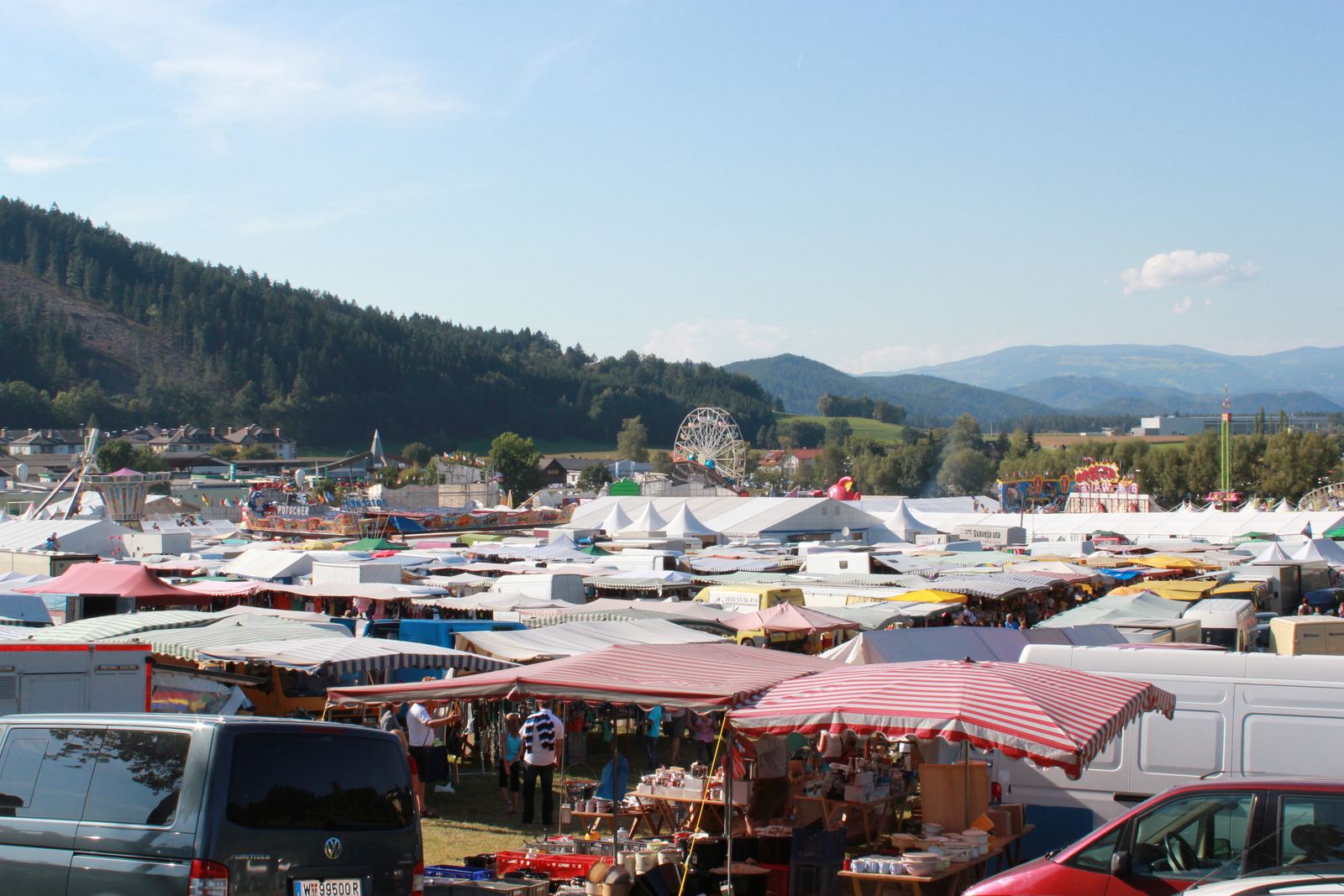 Der Bleiburger Wiesenmarkt: unzählige Marktstände und Zelte unter blauem Himmel, dahinter Riesenrad und Berge