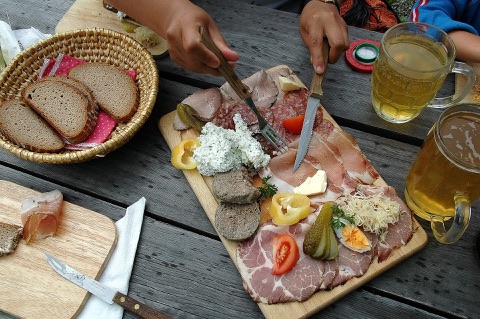 Kärntner Brettljause mit Schinken, Speck, Salami, Topfenaufstrich, Gurkerl und Ei auf einem Holzbrett, daneben ein Brotkorb und zwei Krüge Most auf einem Holztisch