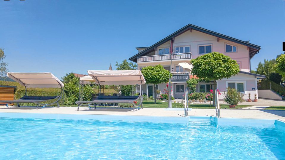 Pool im Vordergrund, dahinter das Ferienhaus mit Balkonen, Doppelliegen und Sonnenschirmen