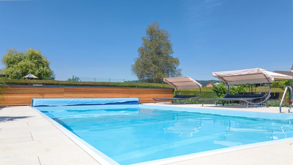 Pool mit aufgerollter Abdeckung, Doppelliegen und Bäume am Rand der Liegewiese