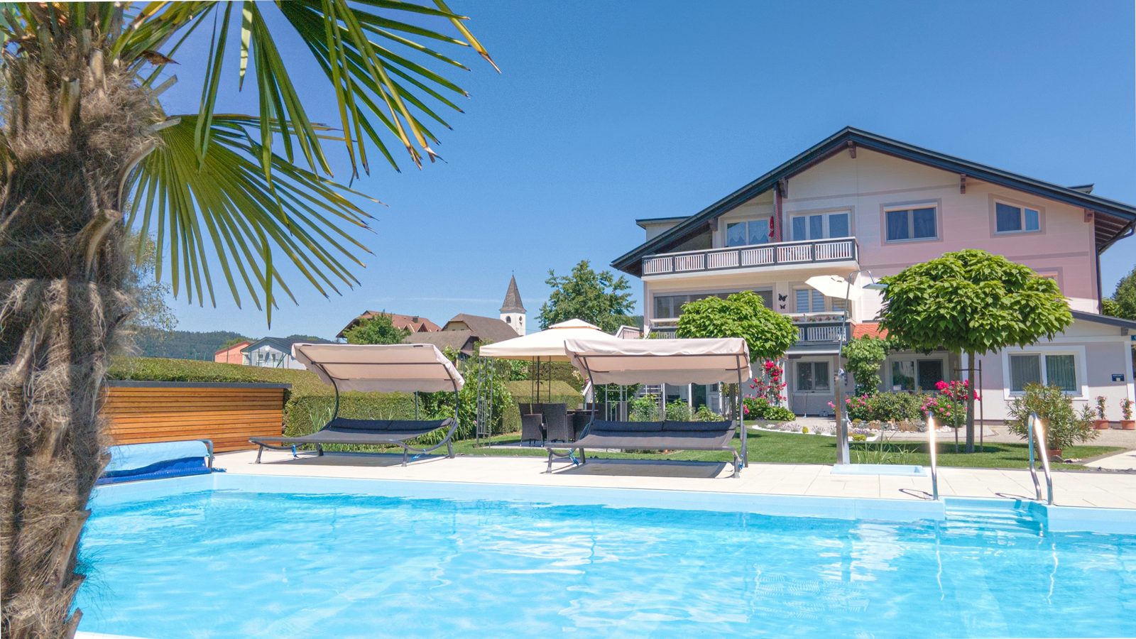 Pool mit Palme im Vordergrund, dahinter Doppelliegen, Sonnenschirm und das Haus mit Balkonen